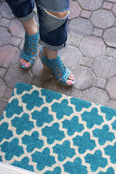 A teal doormat with a crosscross white Moroccon style print with a woman standing next to it.