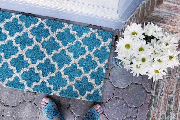 A teal doormat with a crosscross white Moroccon style print and a bouquet of flowers at the corner.