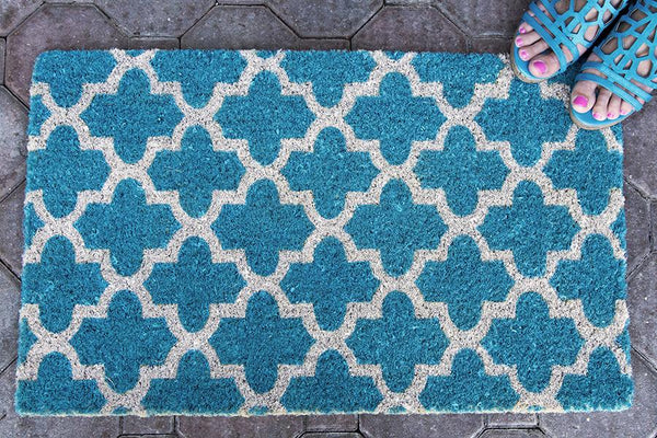 A teal doormat with a crosscross white Moroccon style print with a woman's feet on it in one corner.
